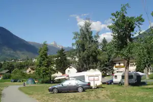 Campingplatz mit Auto, Wohnwagen und Zelten auf einer Wiese, umgeben von Bäumen und Häusern vor den Bergen.