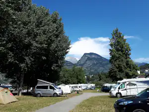 Campingplatz mit Wohnmobilen und Vans am Weg, umgeben von Bäumen, im Hintergrund Berge und ein klarer Himmel.