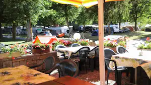 Terrasse am Campingplatz mit Tisch und Stühlen, Blumenbeeten und Sonnenschirm; im Hintergrund Bäume, Fahrzeuge und weitere Stellplätze.