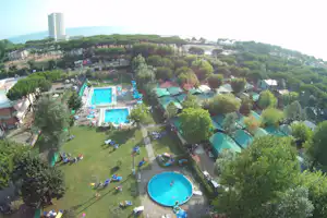 Blick auf Campinganlage mit zwei Schwimmbecken, Liegen und Wegen zwischen Olivenbäumen, im Hintergrund Meer und Hochhaus.