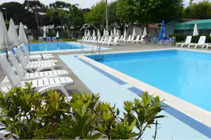 Schwimmbecken mit blauen Bahnen, weißen Liegen und Sonnenschirmen am Campingplatz, im Hintergrund Bäume und Zelte.