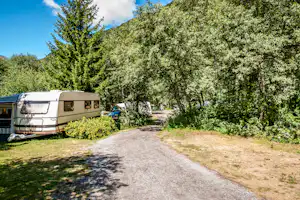 Wohnwagen auf dem Campingplatz, Kiesweg zwischen Bäumen und Hecken, im Hintergrund weitere Fahrzeuge und Zelte.