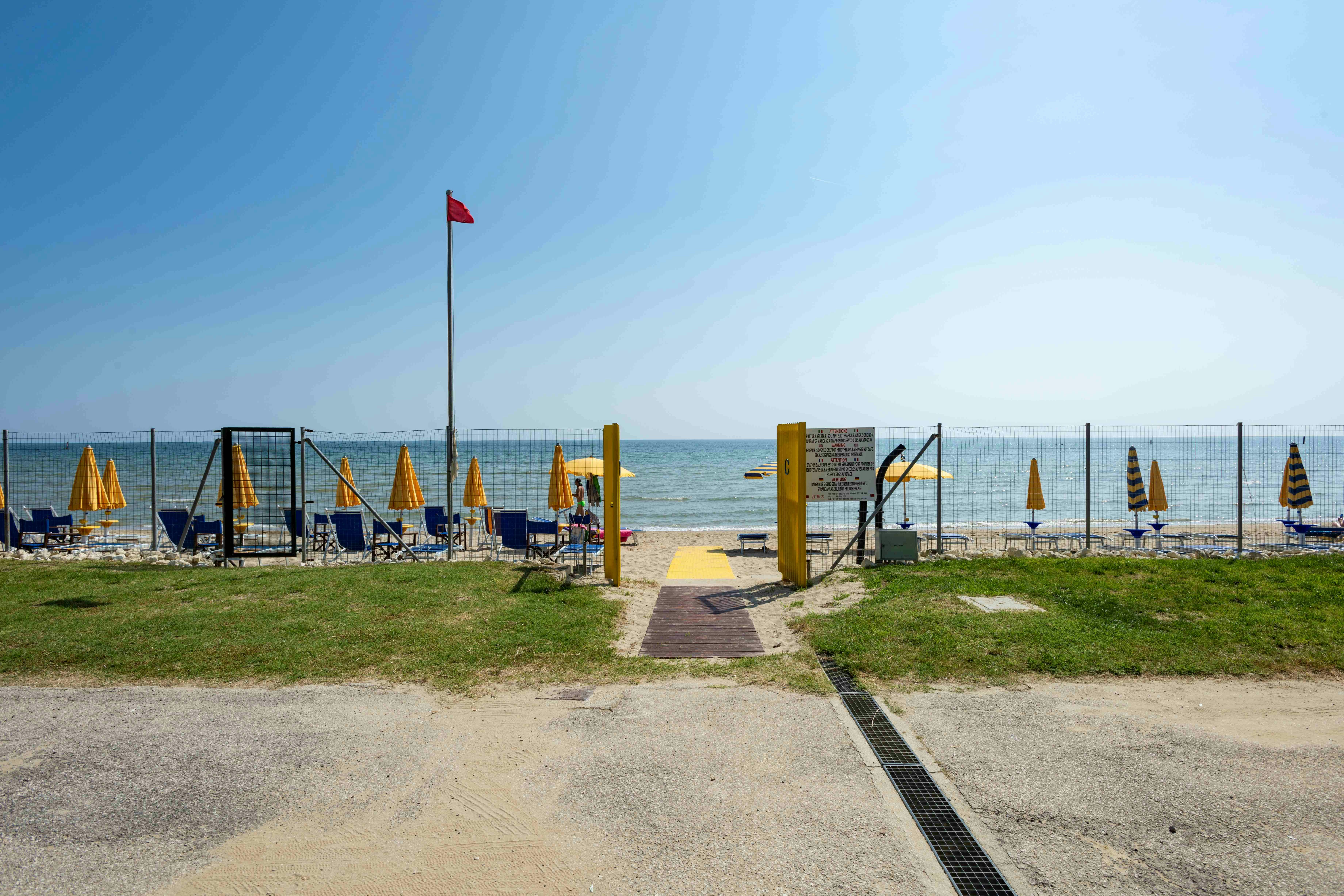 Camping am Strand: Zaun, gelbe Pfosten, Liegen und Sonnenschirme, Weg zum Meer und Blick auf das Wasser.