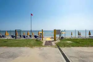 Camping am Strand: Zaun, gelbe Pfosten, Liegen und Sonnenschirme, Weg zum Meer und Blick auf das Wasser.
