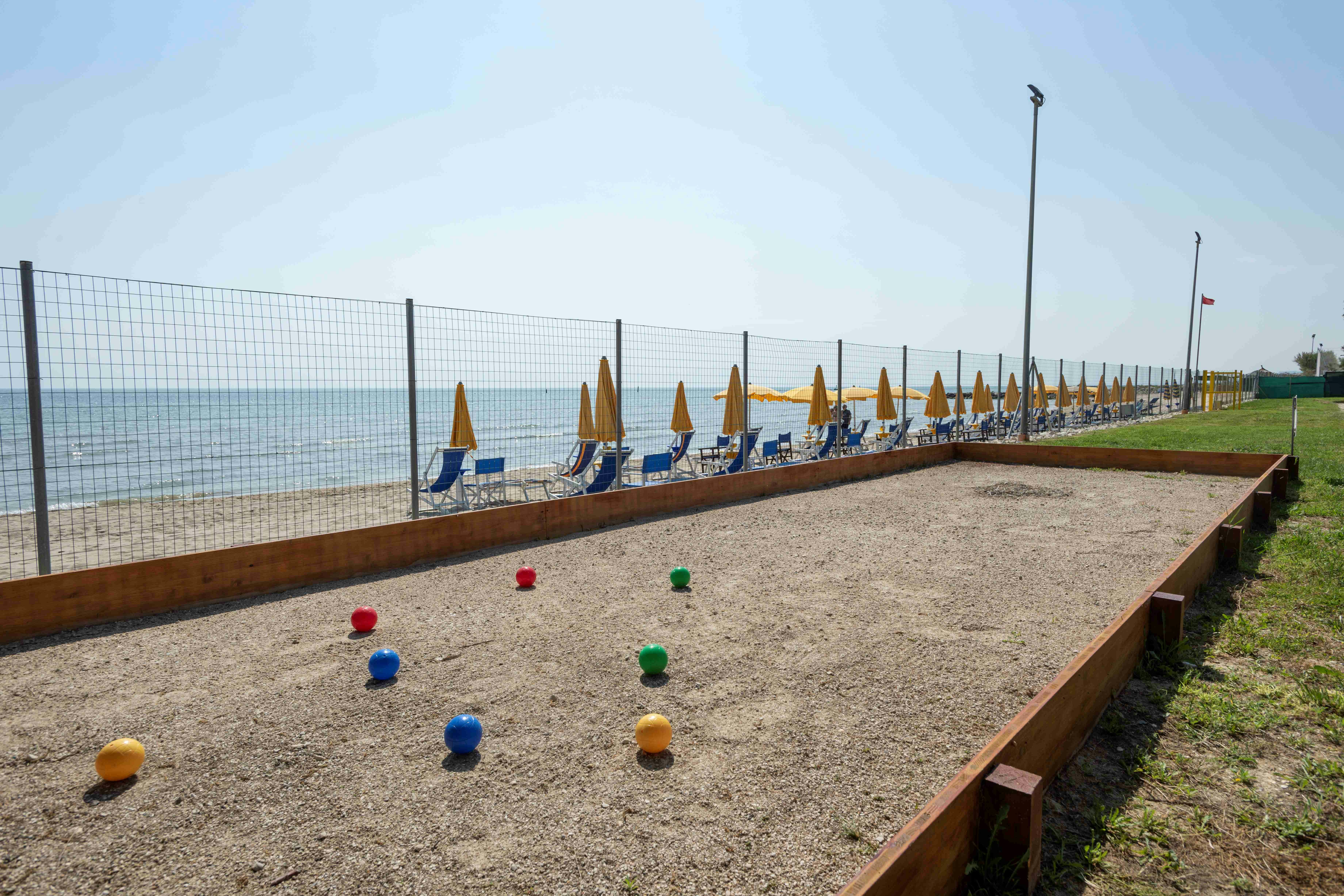 Boulebahn am Campingplatz mit bunten Kugeln, Holzrand und Blick auf das Meer, daneben Sitzbereich und Zaun.