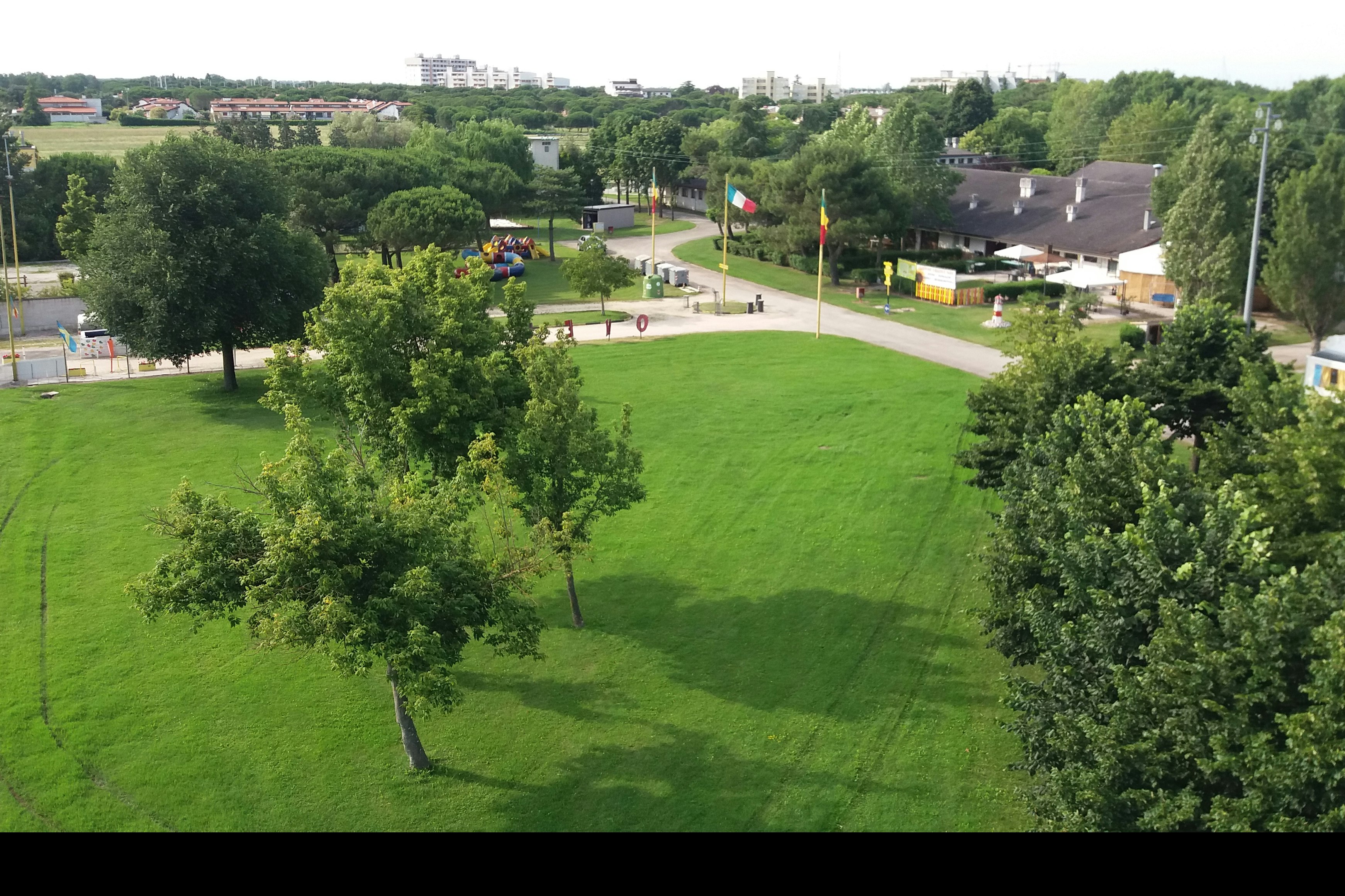 Weite Rasenfläche auf einem Campingplatz mit Wegen, Bäumen und mehreren Wohnwagen, im Hintergrund Häuser und Grün.