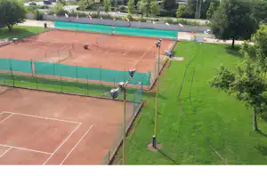 Blick auf einen Tennisplatz mit Umzäunung und Netzen, daneben ein großer Pool und gepflegte Rasenflächen im Campingbereich.