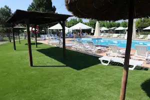 Schwimmbad mit mehreren Liegen auf dem Rasen, Sonnenschirme und ein Holzpavillon im Vordergrund.