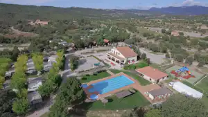 Luftaufnahme einer Campinganlage mit blauem Pool, Wohngebäude, roten Dächern, gepflegten Wegen und viel Grün in hügeliger Landschaft.