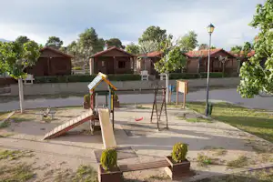 Spielplatz mit Schaukel und Klettergerüst auf dem Campingplatz, im Hintergrund mehrere Holzhütten und Bäume.