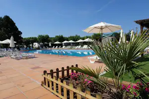 Poolbereich mit Liegen und Sonnenschirmen, gepflasterte Terrasse, Blumenbeet und Holzgeländer im Vordergrund.