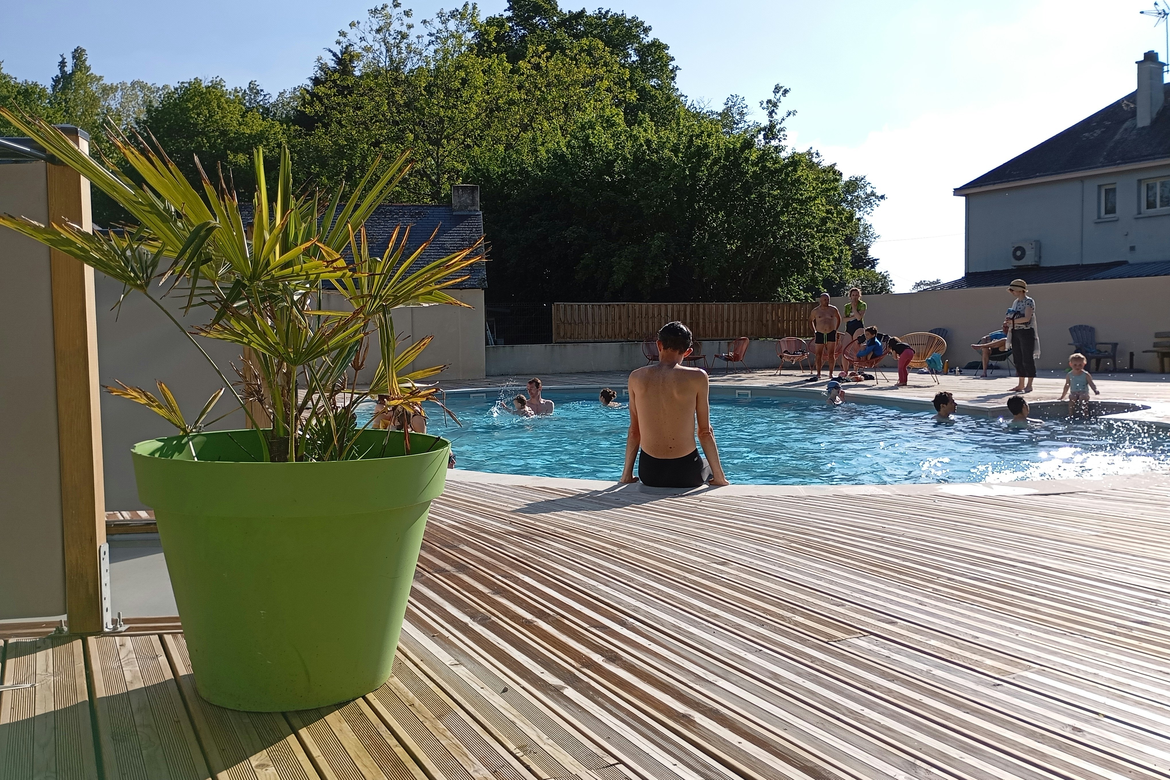 Pool auf dem Campingplatz mit Holzterrasse, grünen Pflanzkübeln und mehreren Badegästen im Hintergrund.
