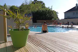 Pool auf dem Campingplatz mit Holzterrasse, grünen Pflanzkübeln und mehreren Badegästen im Hintergrund.