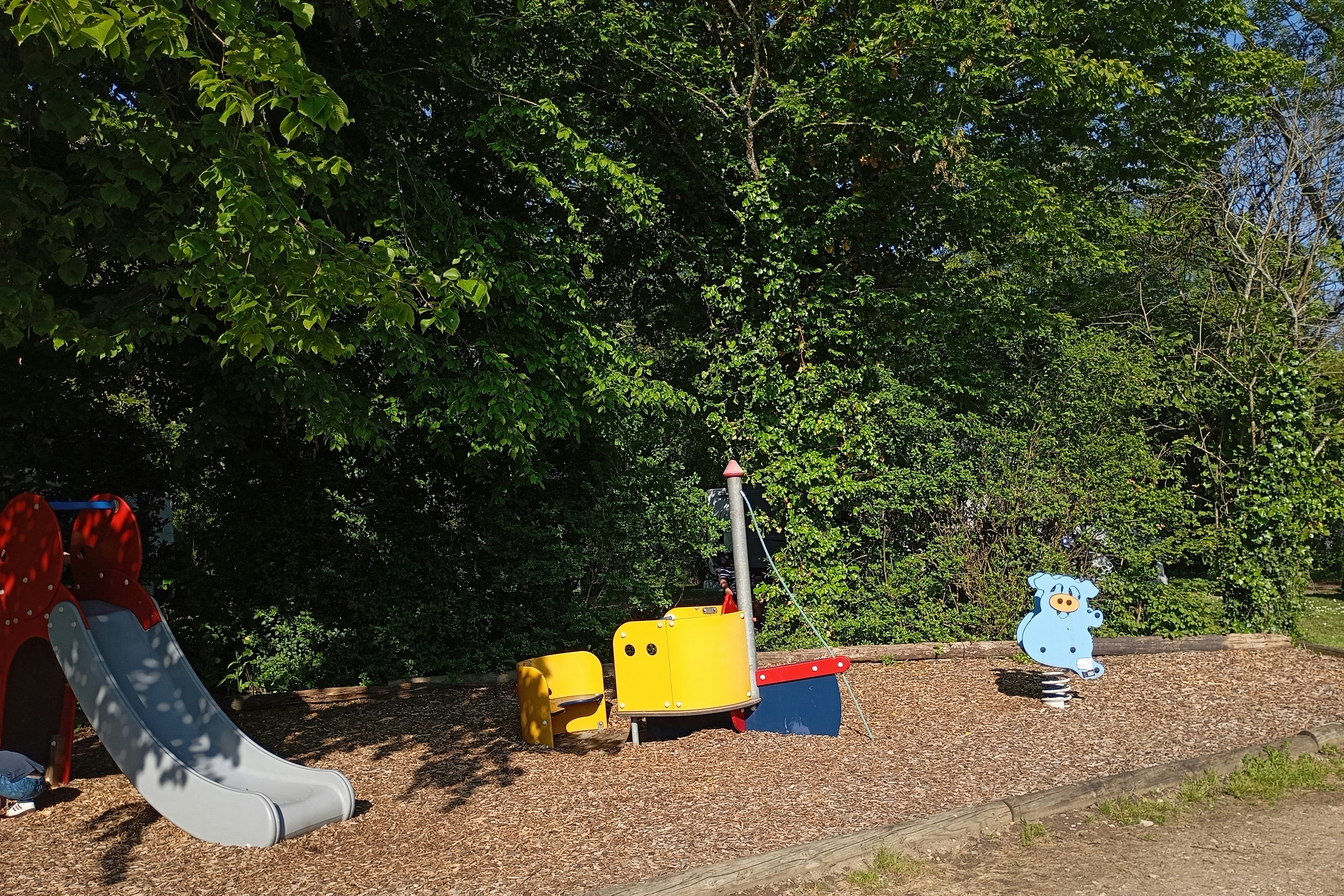 Spielbereich mit gelben Spielgeräten und Rutsche auf Kiesfläche, umgeben von dichtem Grün und Bäumen.
