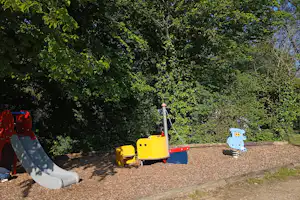 Spielbereich mit gelben Spielgeräten und Rutsche auf Kiesfläche, umgeben von dichtem Grün und Bäumen.
