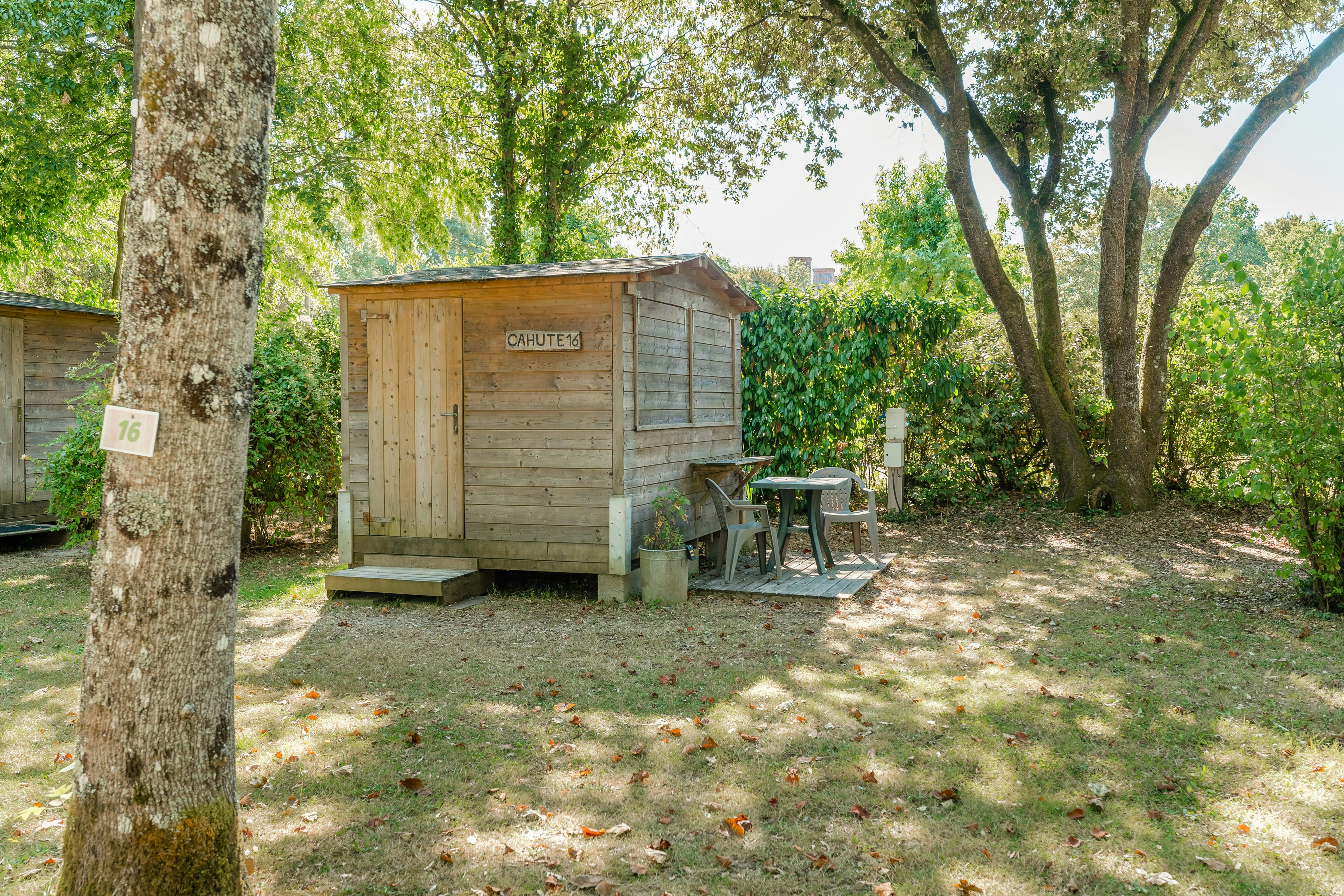 Holzhütte mit Veranda und Gartenmöbeln im Campingbereich, umgeben von Bäumen und Rasenfläche.