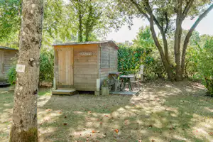 Holzhütte mit Veranda und Gartenmöbeln im Campingbereich, umgeben von Bäumen und Rasenfläche.
