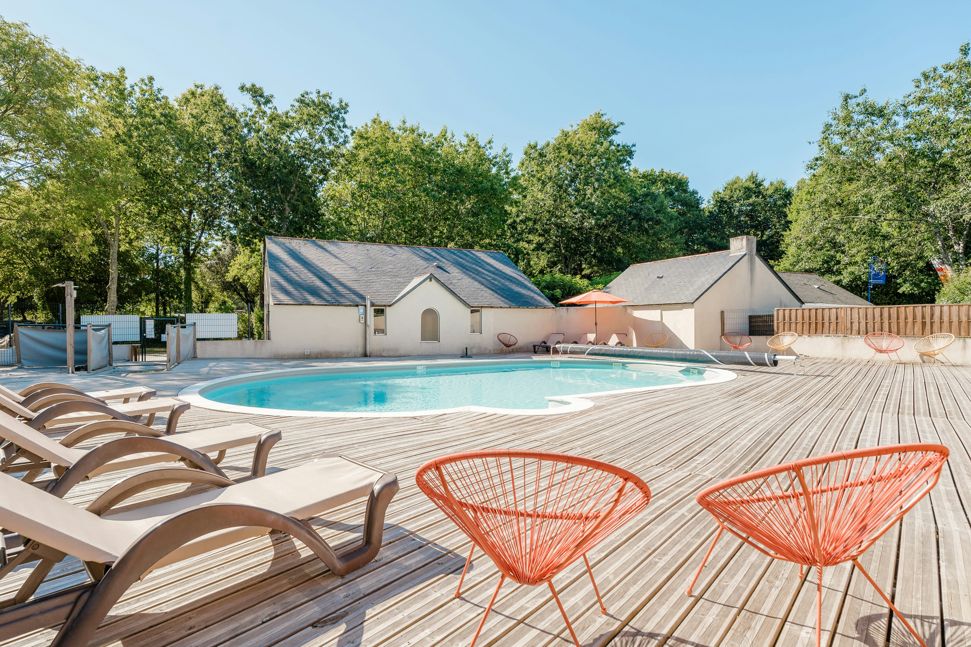 Poolbereich mit Holzdeck, orangefarbenen Stühlen und Sonnenliegen vor einem Campingplatzgebäude, umgeben von Bäumen.