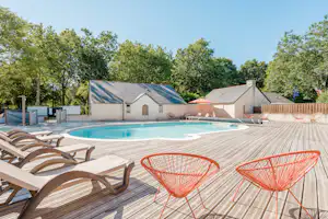 Poolbereich mit Holzdeck, orangefarbenen Stühlen und Sonnenliegen vor einem Campingplatzgebäude, umgeben von Bäumen.