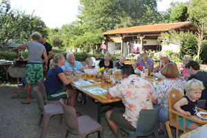 Familie sitzt an einem langen Holztisch im Campinggarten, im Hintergrund ein überdachter Bereich und ein Wohnmobil.