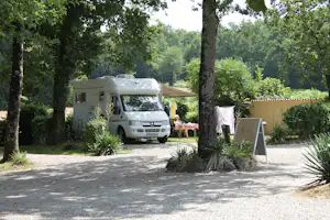 Wohnmobil auf einem Campingplatz zwischen Bäumen, mit Tisch und Sonnenschirm am Stellplatz.