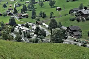 Luftaufnahme eines Campingplatzes mit Wohnwagen, kleinen Hütten und Gebäuden in grünen Wiesen, umgeben von Bäumen und Wegen.