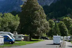 Wohnmobile auf dem Campingplatz, im Hintergrund Häuser und grüne Berge, umrahmt von Bäumen und Talwiesen.
