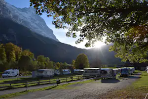 Campingplatz mit Wohnmobilen und Caravans, Blick auf hohe Berge, Bäume im Vordergrund und Sonnenlicht am Talhang.