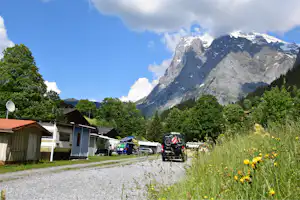 Campingplatz mit Wohnmobilen und Wohnwagen am Weg, umgeben von Wiesen und Bäumen, im Hintergrund hohe Alpenberge.
