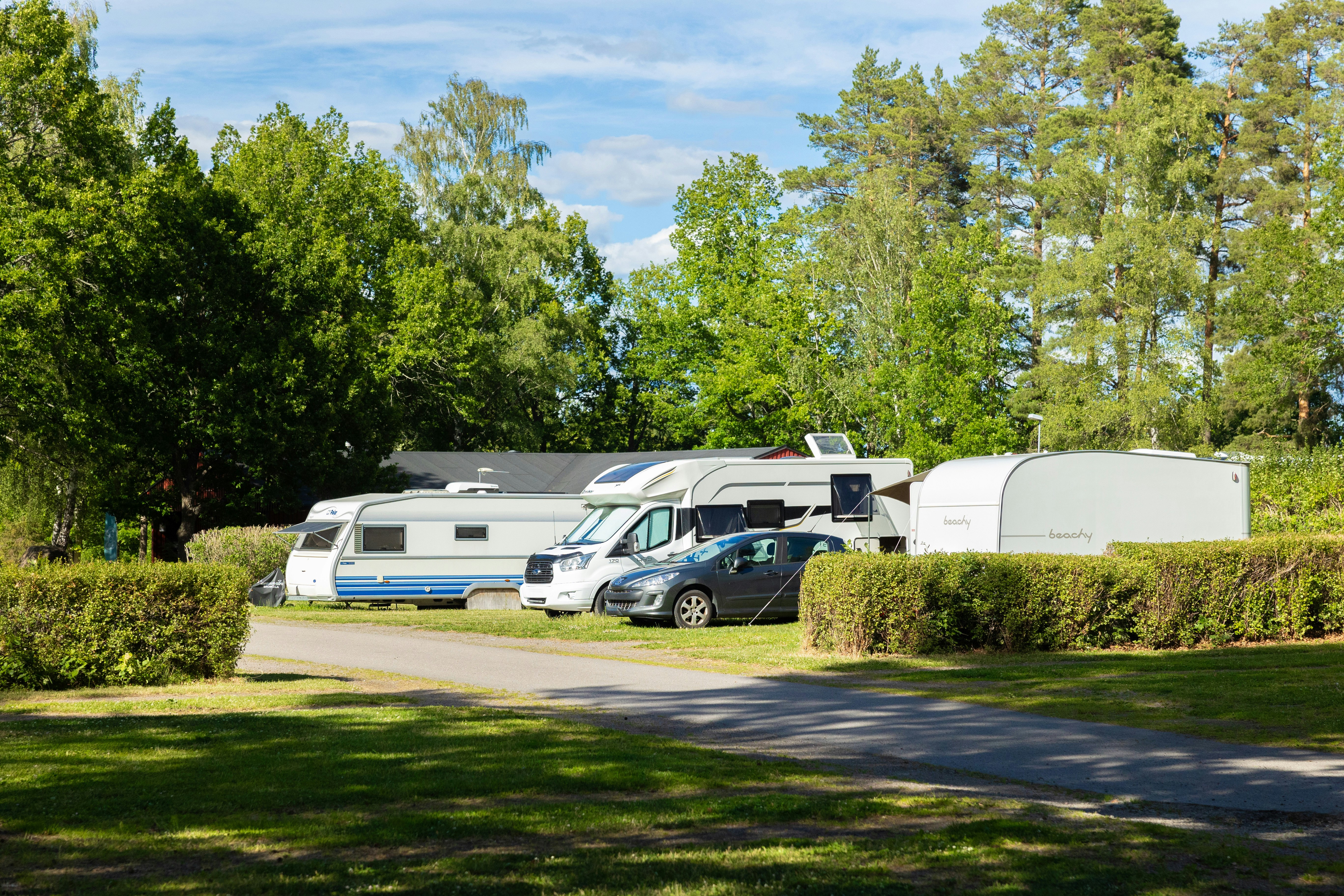 Wohnmobile und ein Campingbus stehen auf einem Stellplatz zwischen Hecken und Bäumen, im Hintergrund ein weiteres Fahrzeug.