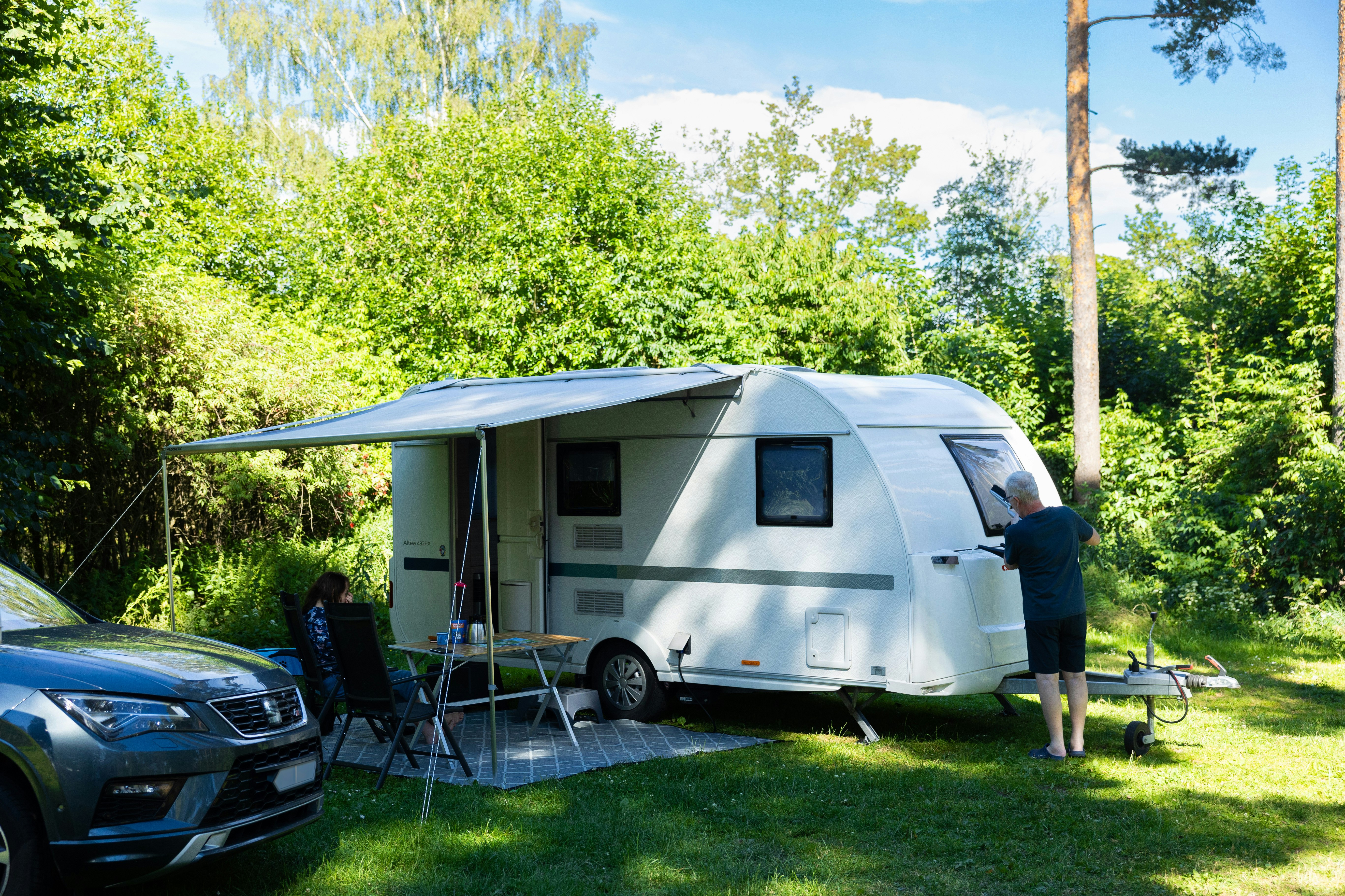 Wohnwagen mit Vorzelt auf dem Campingplatz, daneben Auto und Tisch, im Hintergrund Bäume und grünes Gras.