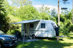 Wohnwagen mit Vorzelt auf dem Campingplatz, daneben Auto und Tisch, im Hintergrund Bäume und grünes Gras.