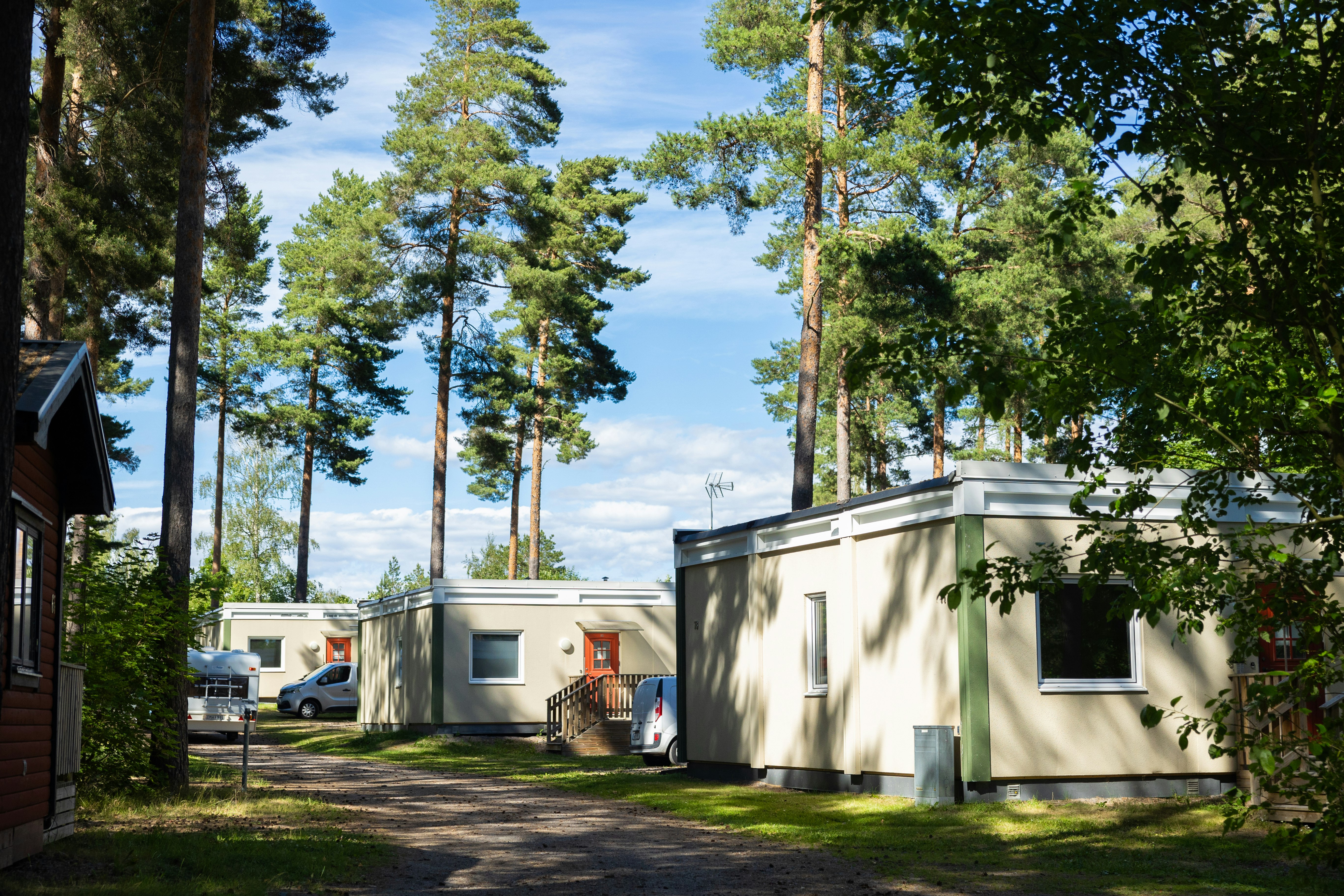 Stellplätze im Campingwald mit Wohnwagen und Wohnmobil, weißem Gebäude und Bäumen im Hintergrund.