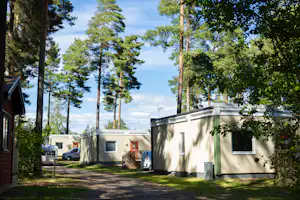 Stellplätze im Campingwald mit Wohnwagen und Wohnmobil, weißem Gebäude und Bäumen im Hintergrund.