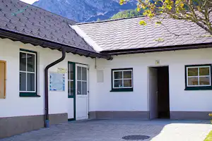 Weiße Ferienhaus-Fassade mit Terrasse, Türen und Fenstern, davor gepflasterter Bereich und im Hintergrund Berge.