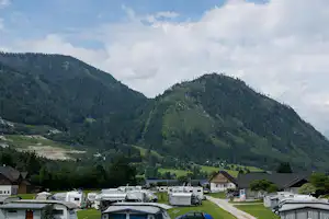 Campingplatz im Tal mit Wohnmobilen und Wohnwagen, gepflastertem Weg und Häusern am Hang unter grünen Bergen.