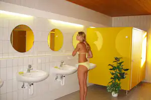 Frau im Bikini vor Waschbecken und Duschbereich mit gelben Türen, Fliesenwand und Pflanzenkübel im Sanitärraum.