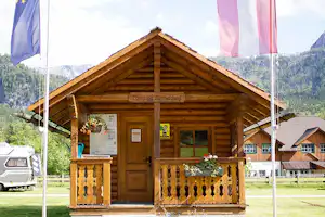 Holz-Hütte am Campingplatz mit Eingangstür, Balkonbrüstung und Blumen vor dem Gebäude, im Hintergrund Berge und Wohnwagen.