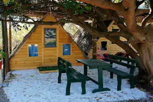 Holzhütte mit großen Fenstern, davor grüne Holzbänke und Hängematte unter einem großen Baum im Campingbereich.