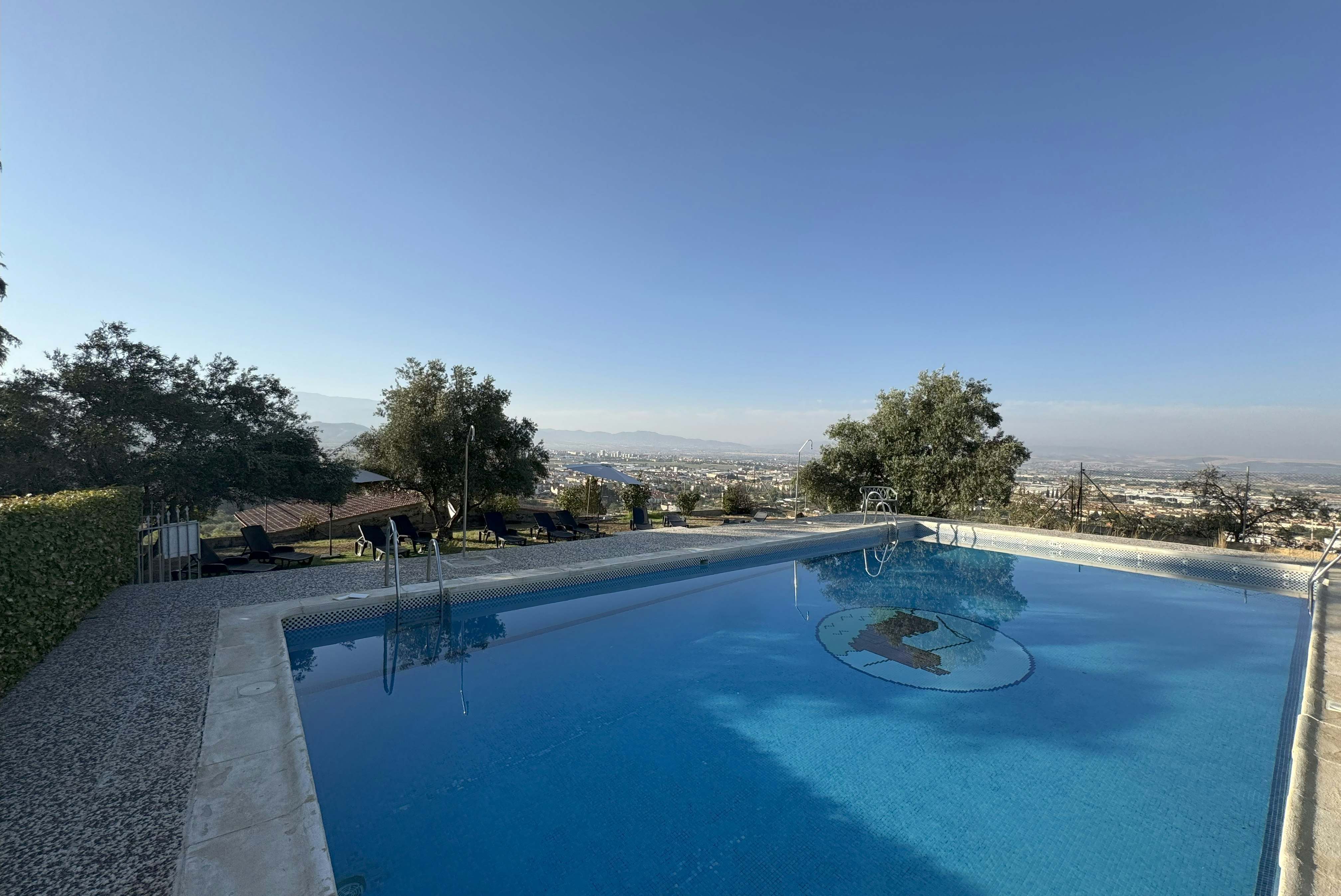 Blaue Camping-Poolanlage mit Liegestühlen und Blick ins Tal, umgeben von Bäumen und Kiesweg am Rand.