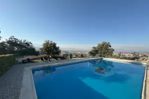 Blaue Camping-Poolanlage mit Liegestühlen und Blick ins Tal, umgeben von Bäumen und Kiesweg am Rand.