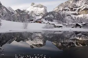 Winterliche Berglandschaft mit Dorf am Seeufer, verschneite Häuser und spiegelnde Reflexionen im ruhigen Wasser.
