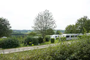 Wohnmobile auf einem Campingplatz im Grünen, mit Hecken, Kiesweg und Blick auf die hügelige Landschaft.