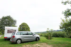Stellplatz am Campingplatz mit silbernem Auto und Wohnwagen neben einem Grasbereich und Bäumen.