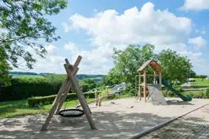 Spielplatz auf dem Campingplatz mit Sandfläche, Rutsche und Klettergerüst, umgeben von Bäumen und Wiesen.