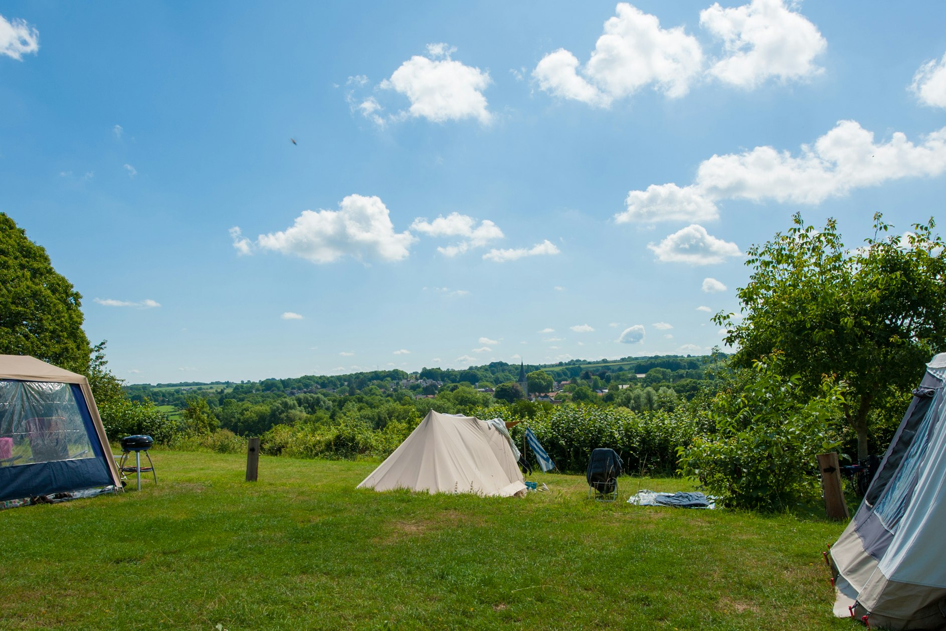 Campingwiese mit mehreren Zelten, grünen Bäumen und einem Weg, im Hintergrund eine weite Landschaft unter blauem Himmel.