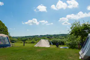 Campingwiese mit mehreren Zelten, grünen Bäumen und einem Weg, im Hintergrund eine weite Landschaft unter blauem Himmel.