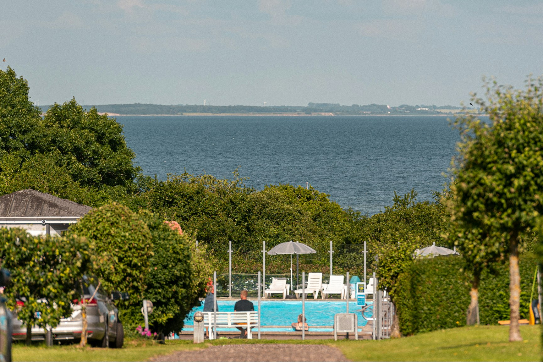 Blick auf einen Campingbereich mit Pool und Liegen, umgeben von Hecken und Bäumen, im Hintergrund das Meer.