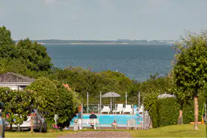 Blick auf einen Campingbereich mit Pool und Liegen, umgeben von Hecken und Bäumen, im Hintergrund das Meer.