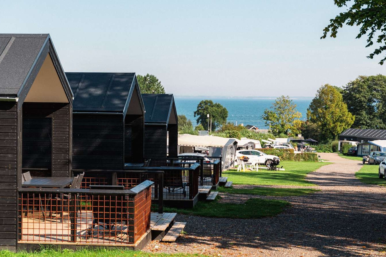 Holzhütten und Stellplätze am Campingplatz, Blick auf das Meer, Rasen und Bäume entlang des Weges.